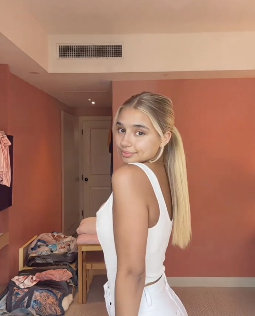 An AI-generated portrait of a young blonde woman in a white dress, posed in a styled hotel-room setting, an example of the synthetic creator-style imagery now scaling on YouTube faceless channels and Shorts. The image is realistic at a glance but reflects the kind of consistent-lighting, single-subject content covered in this post's detection signs.